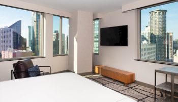 A modern hotel room with a large bed, wall-mounted TV, armchair, and views of city skyscrapers through multiple windows.