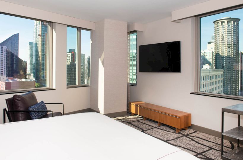 A modern hotel room with a large bed, wall-mounted TV, armchair, and views of city skyscrapers through multiple windows.