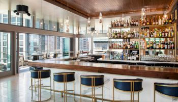 This image shows a stylish bar with high-back chairs, a countertop, shelves of drinks, large windows, and elegant hanging lights.