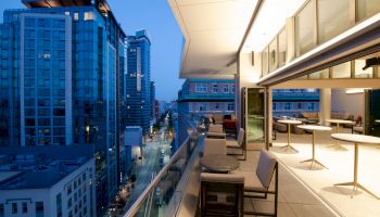 A modern rooftop terrace at dusk, overlooking a city street with tall buildings, featuring tables and chairs for outdoor seating.