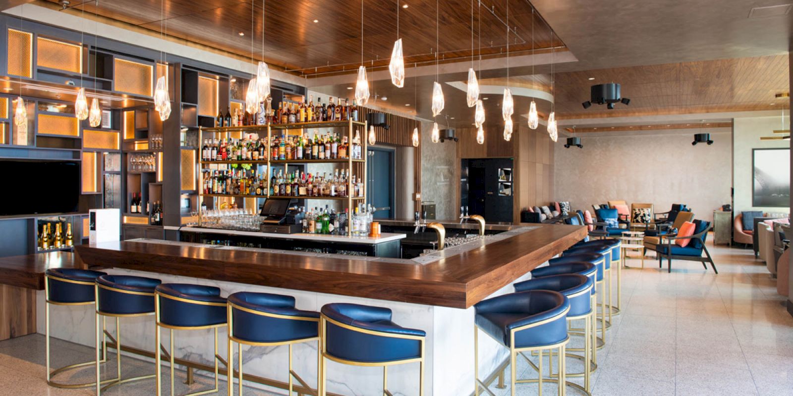 A modern bar with blue stools, hanging lights, and a wide selection of bottles on shelves, set in a stylish lounge atmosphere.