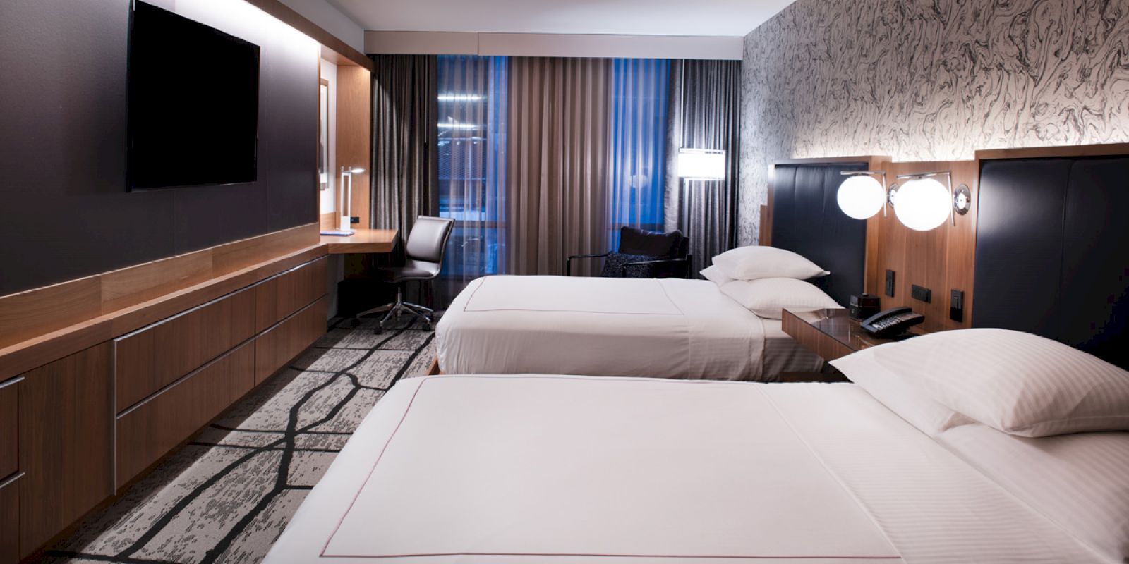 The image shows a modern hotel room with two beds, a flat-screen TV, a desk, and stylish decor including patterned wallpaper and curtains.