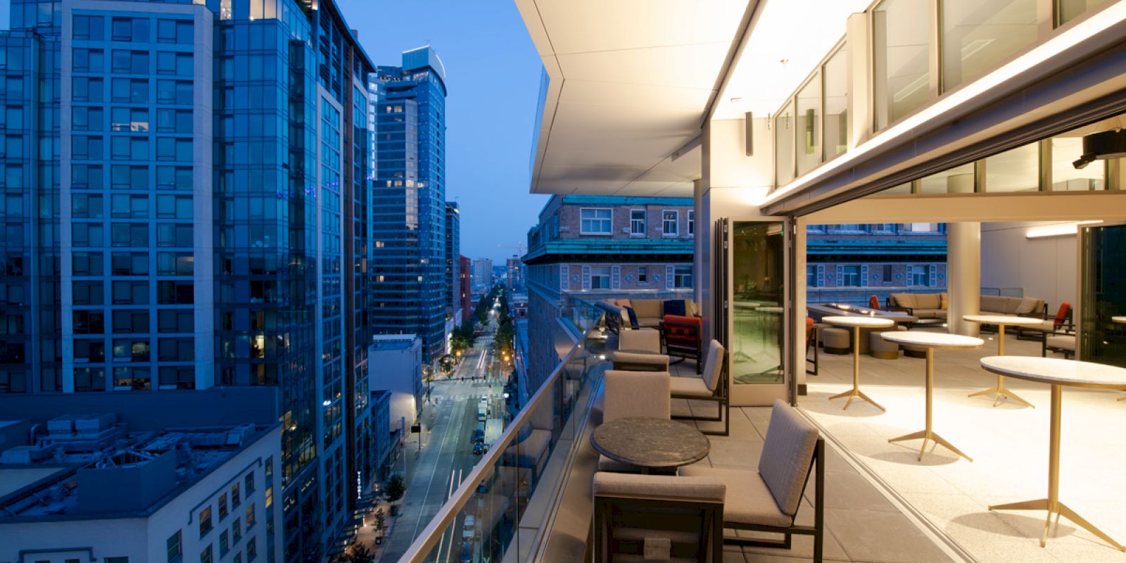 A modern rooftop terrace overlooks a cityscape at dusk, with comfortable seating and large windows glowing with interior light.