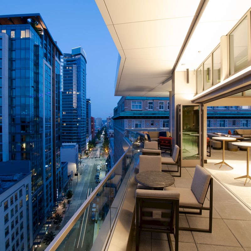 A modern balcony overlooking a cityscape at dusk, featuring outdoor seating and tables, with tall buildings in the background.