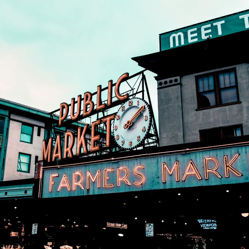 The image shows the iconic neon signs of a public market and farmers market, with a clock, against the backdrop of surrounding buildings.