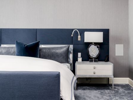 A modern bedroom with a blue headboard, white bedding, a bedside table, lamp, and decorative stone, all on a plush carpet.