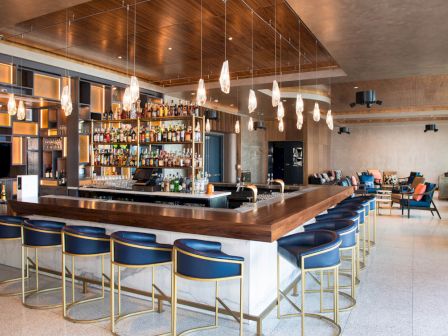 A modern bar with wooden accents, stylish blue chairs, and hanging lights, featuring a well-stocked liquor shelf and cozy seating area.