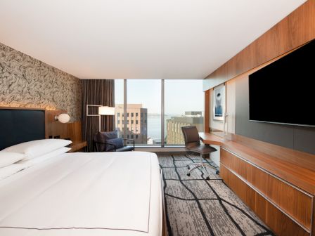 The image shows a modern hotel room with a large bed, desk, chair, TV, and a window view of buildings and water.