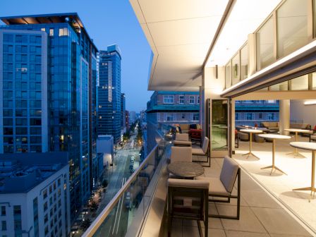 A modern rooftop terrace at dusk, overlooking a city street with tall buildings, featuring outdoor seating and open indoor space.