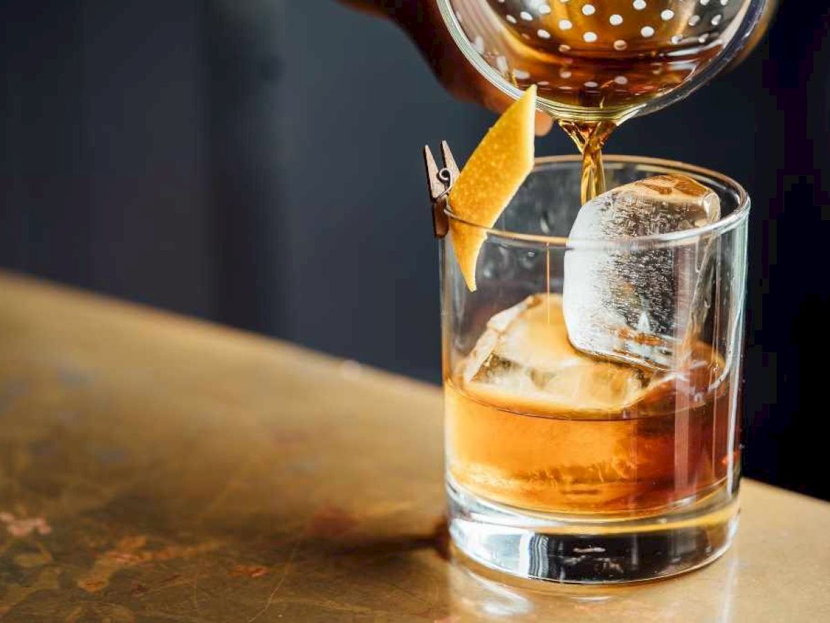 A cocktail is being poured into a glass with ice and a citrus peel garnish, set on a wooden surface.