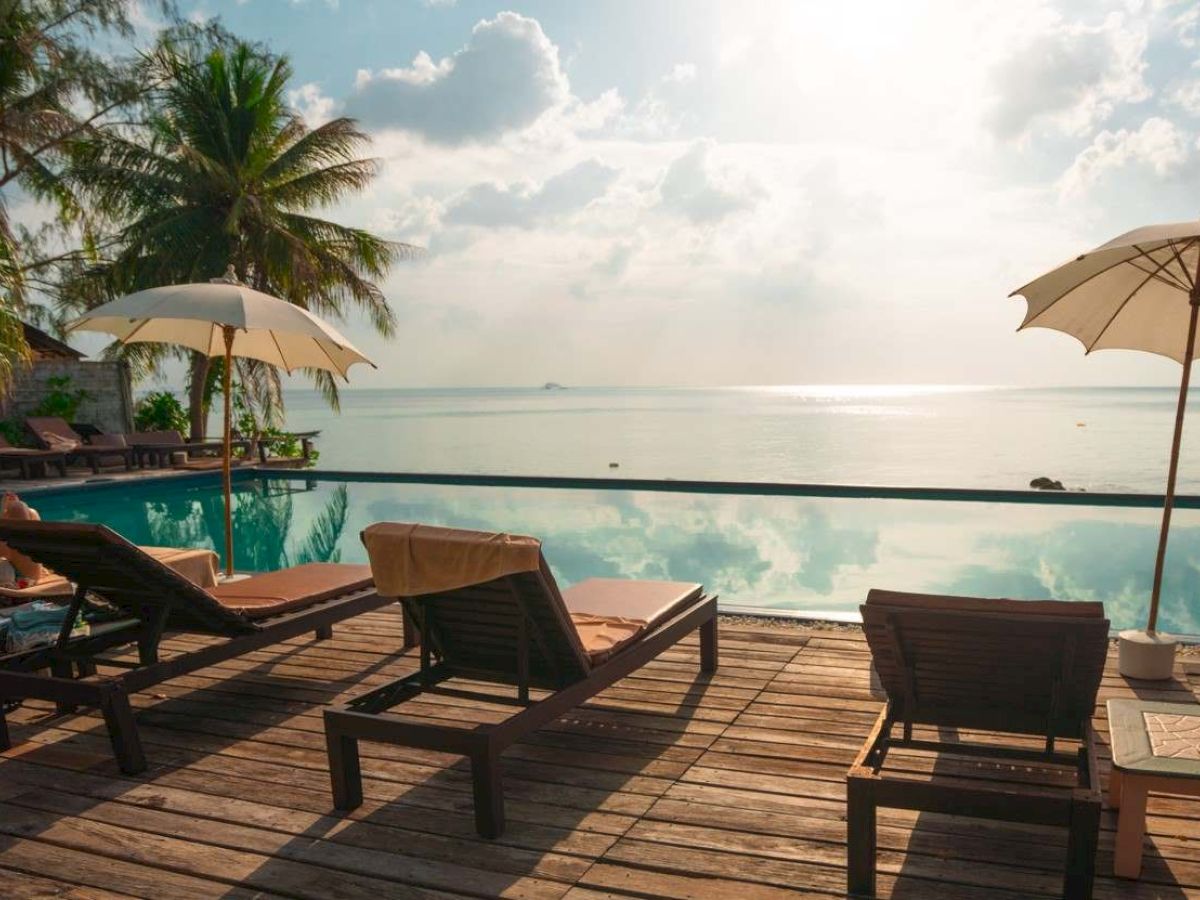A serene tropical setting with lounge chairs and umbrellas by an infinity pool, overlooking a calm sea and palm trees under a sunny sky.