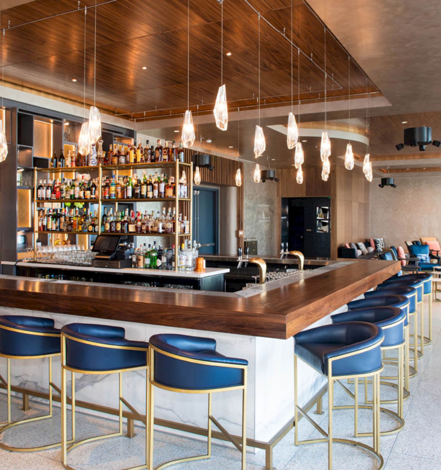 The image shows a stylish bar interior with a large counter, blue chairs, and hanging lights, featuring a fully stocked shelf in the background.