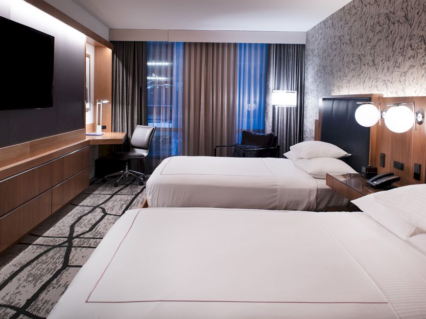 The image shows a modern hotel room with two neatly made beds, a desk, a TV, and stylish decor, featuring a patterned carpet.