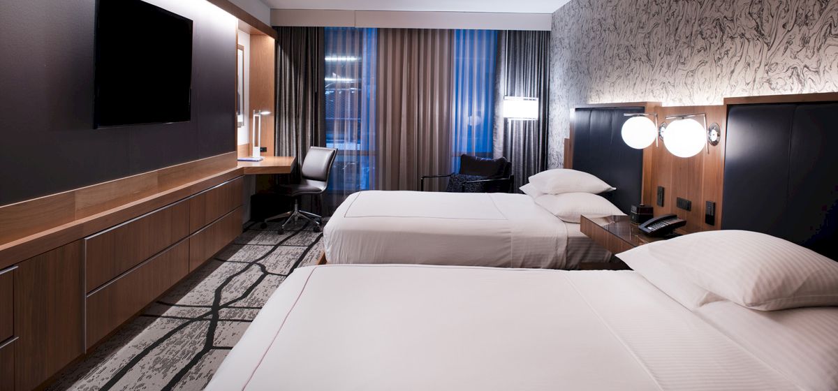 The image shows a modern hotel room with two neatly made beds, a desk, a TV, and stylish decor, featuring a patterned carpet.