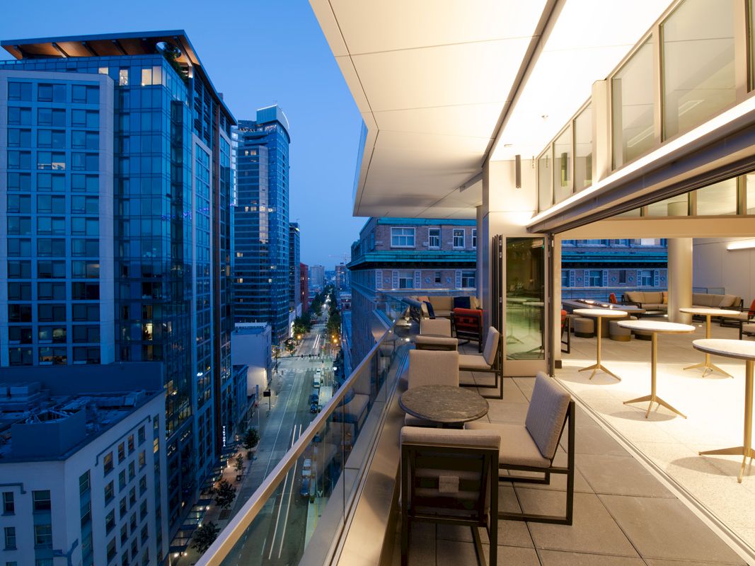 A modern cityscape at twilight with high-rise buildings, a sleek balcony, outdoor seating, and bright lights inside.