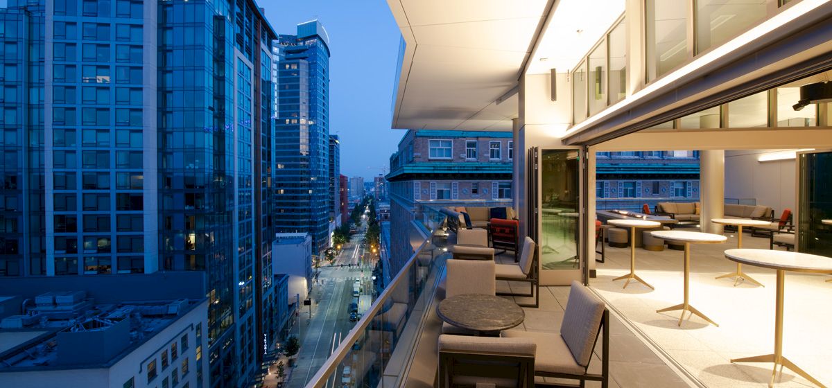 A modern cityscape at twilight with high-rise buildings, a sleek balcony, outdoor seating, and bright lights inside.
