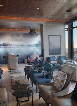 The image shows a modern lounge with elegant seating, large windows with a sea view, and a mural of a cityscape on the wall.