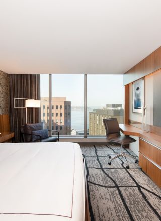 A modern hotel room with a large bed, wall-mounted TV, desk, and a window overlooking city buildings.