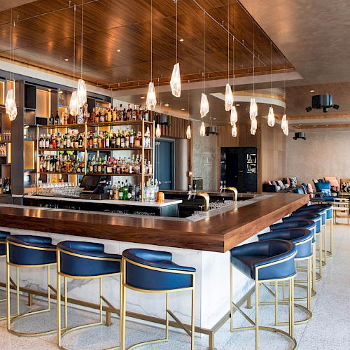 A modern bar with blue stools, hanging lights, and a stocked backbar sits in an open lounge space with seating areas and light decor.