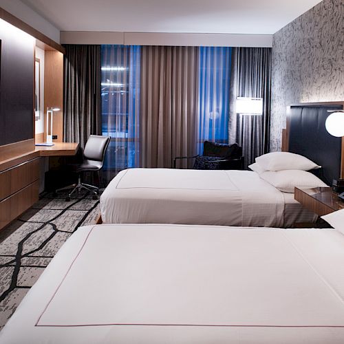A modern hotel room with two beds, a flat-screen TV, a desk with a chair, and warm lighting against patterned wallpaper completes the decor.