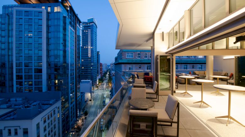 A modern cityscape at dusk with tall buildings and an elegantly furnished balcony overlooking the street.