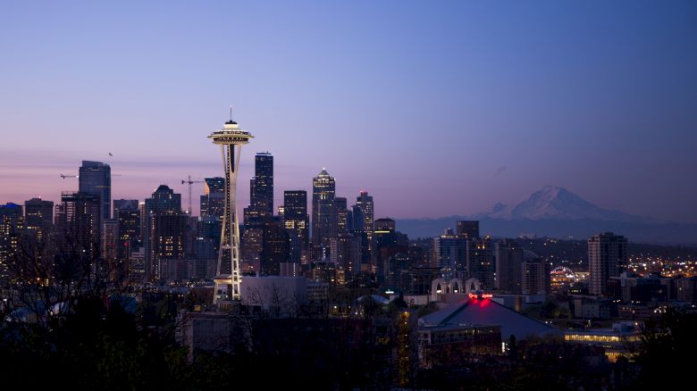 The image shows a city skyline with the Space Needle, skyscrapers, and Mount Rainier in the background during twilight.