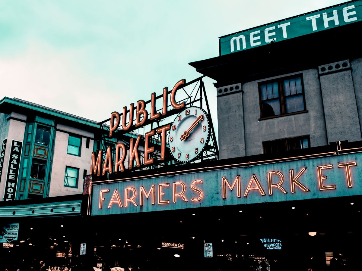 The image shows signs for a "Public Market" and "Farmers Market" with a clock, on a building under cloudy skies.