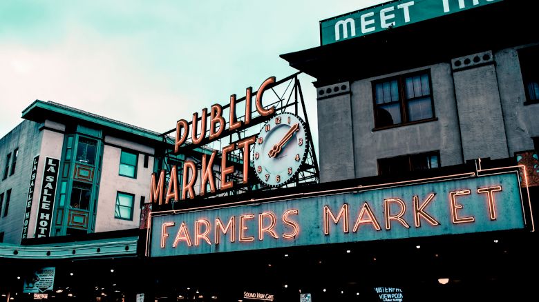 The image shows signs for a "Public Market" and "Farmers Market" with a clock, on a building under cloudy skies.