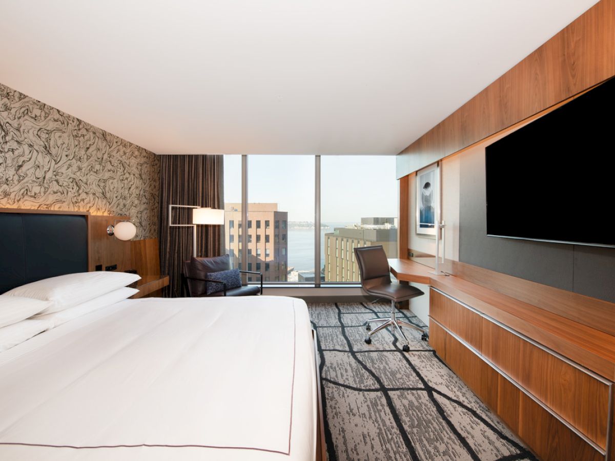 A modern hotel room with a large bed, wall-mounted TV, desk, and a window view of buildings outside.