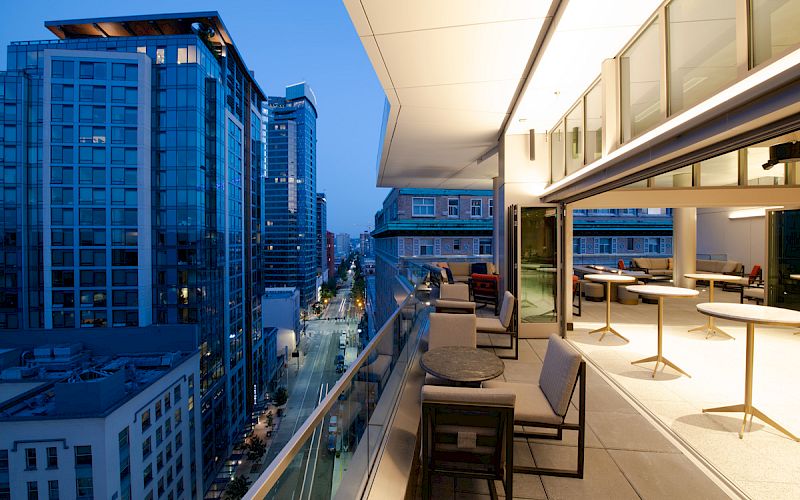 A modern urban balcony view at dusk, featuring a sleek outdoor seating area and overlooking a city street with tall buildings.