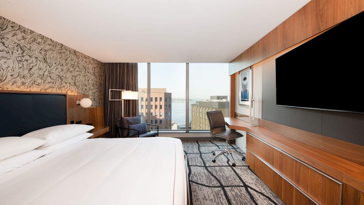 A modern hotel room with a large bed, wall-mounted TV, desk, chair, and window view of city buildings and water.