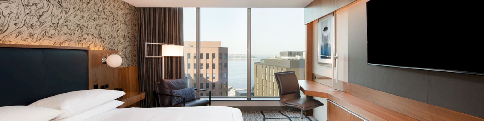 A modern hotel room with a large bed, wall-mounted TV, desk, chair, and window view of city buildings and water.