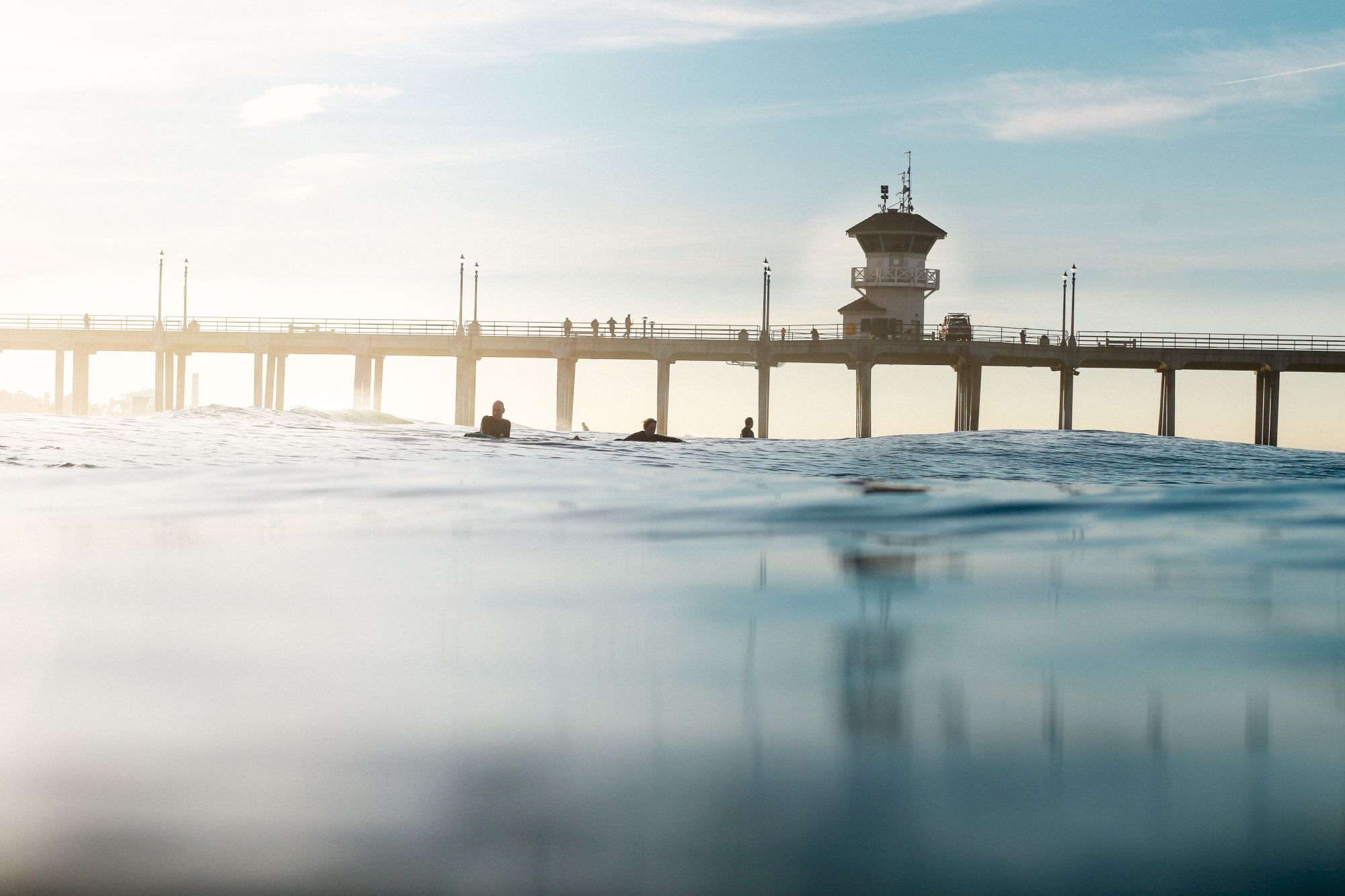 The image shows a pier extending over the ocean with a few people on it and surfers in the water, under a sunny, partly cloudy sky.