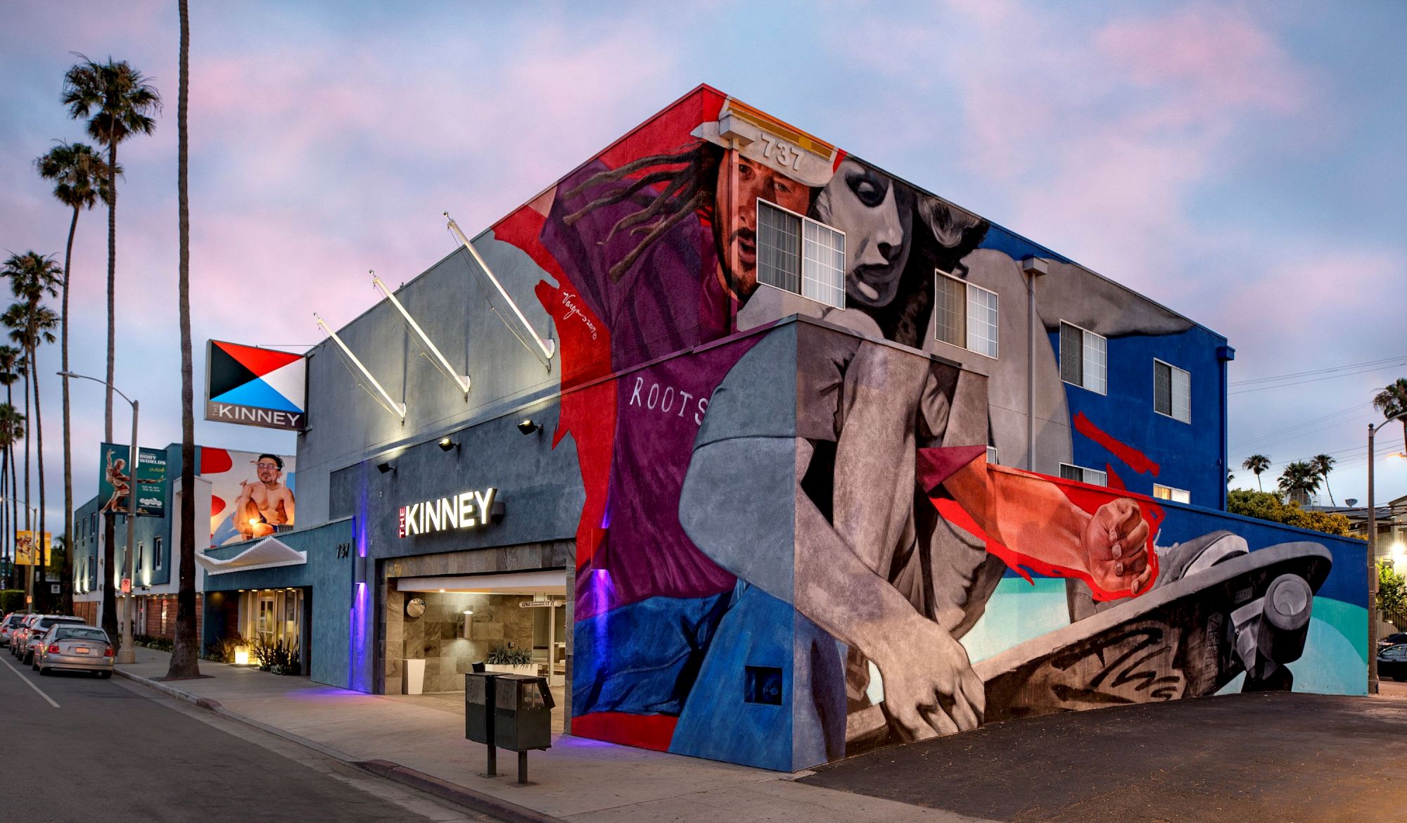 The image shows a vibrant mural on a building housing "The Kinney," featuring dynamic street art with a skateboard theme.
