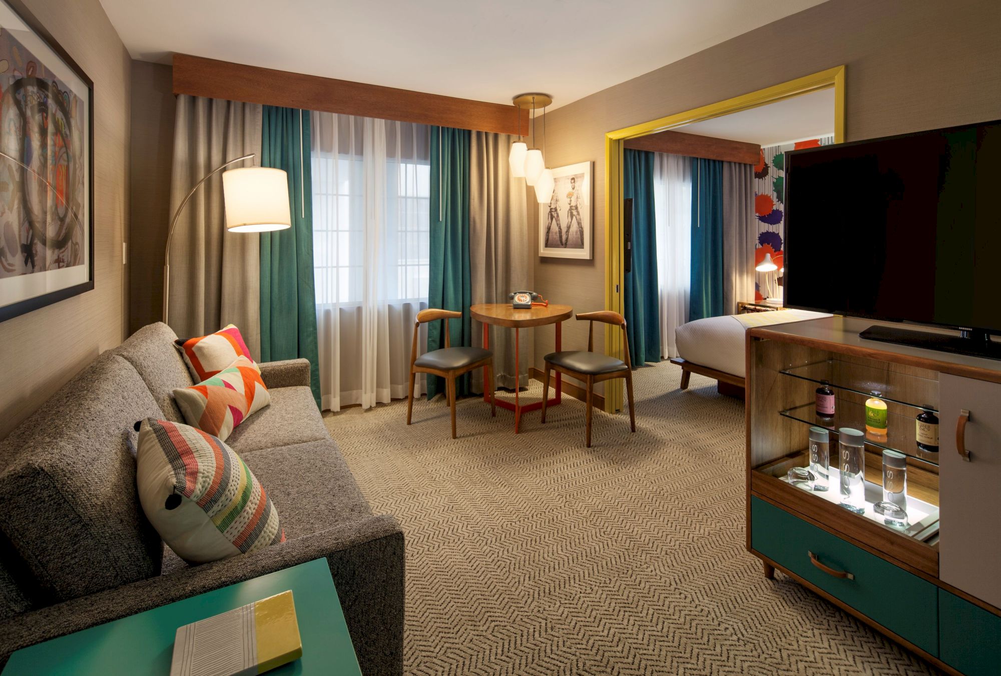 A modern hotel room with a sofa, colorful cushions, a TV, patterned carpet, table with chairs, and a glimpse into a bedroom.