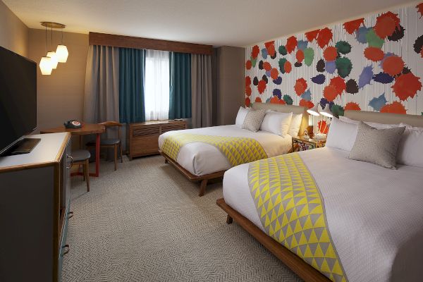 A colorful hotel room with two beds, geometric bed runners, vibrant wall design, desk, chair, TV, and pendant lighting.
