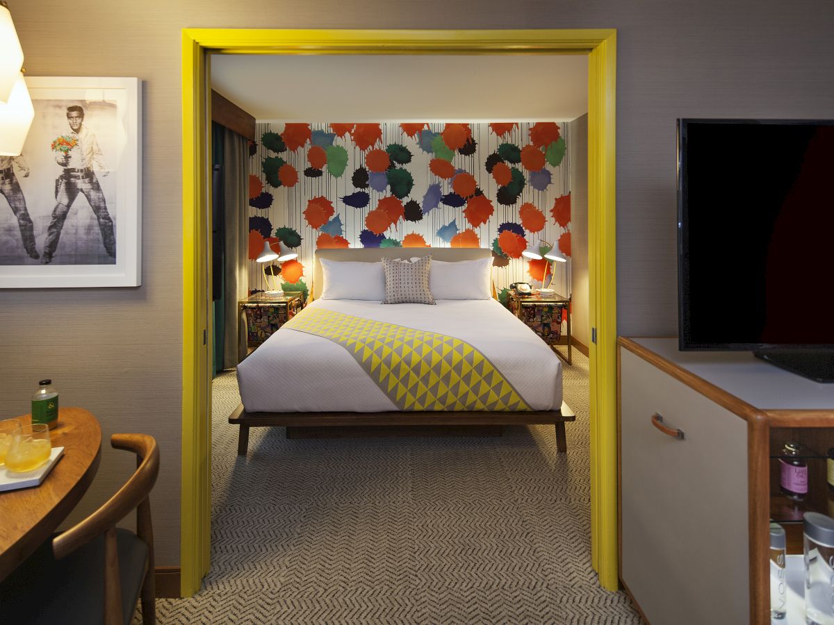 The image shows a stylish hotel room with a vibrant, colorful wall behind a bed, a TV, a minibar, artwork, and warm lighting accents.