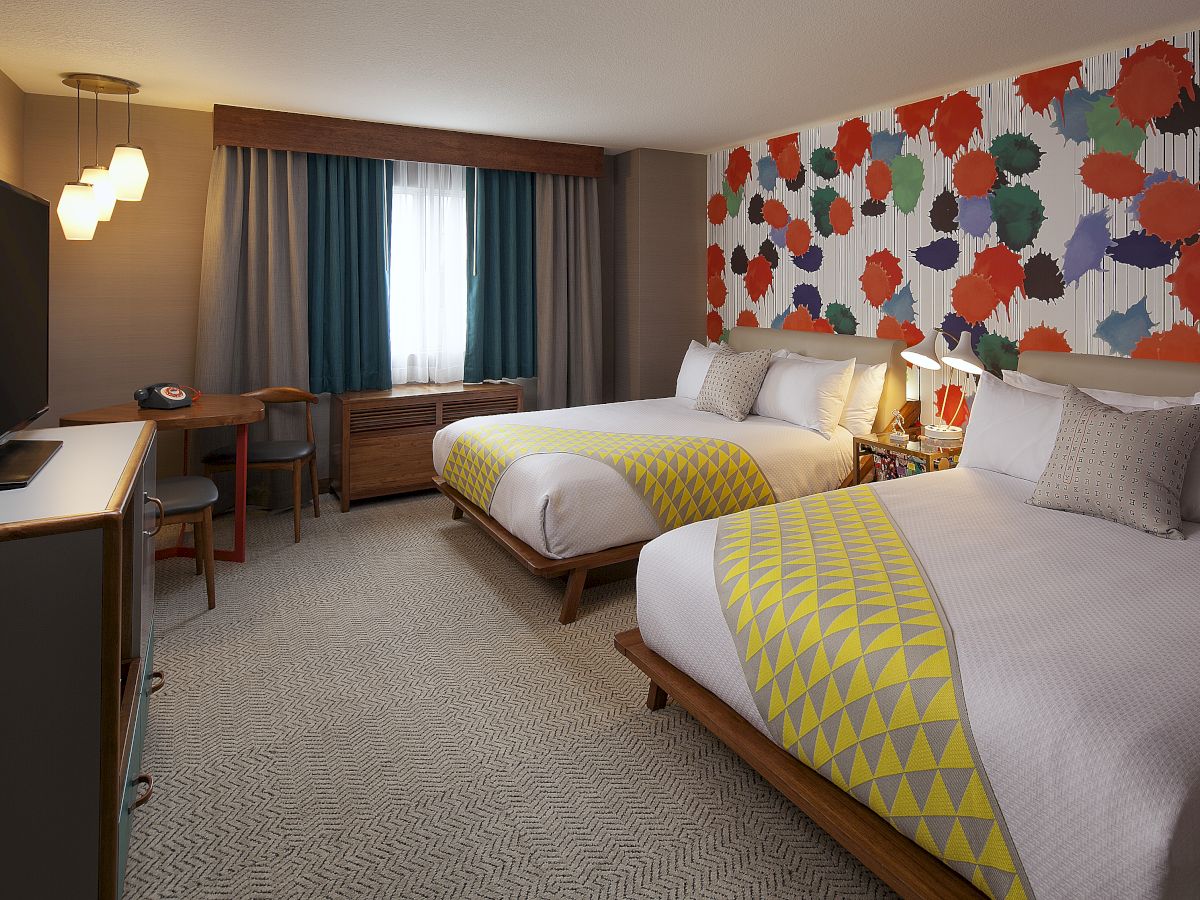 The image shows a hotel room with two beds, colorful wall art, a TV, and a desk near a window with curtains.