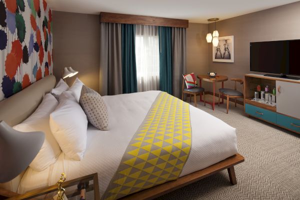 A stylish hotel room with a colorful accent wall, modern furnishings, a bed with geometric bedding, a desk, and a flat-screen TV.