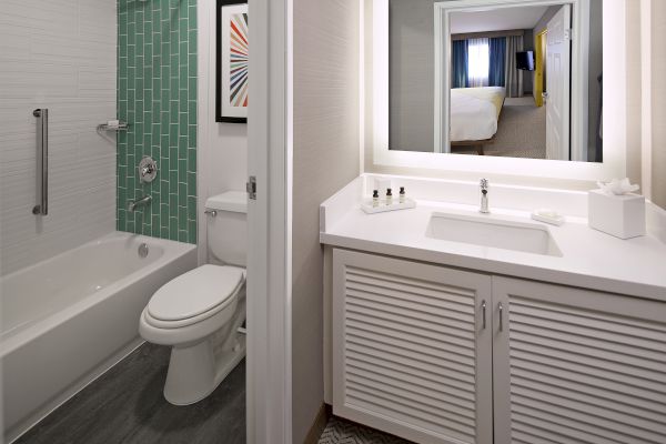 This is a modern bathroom with a bathtub, green tile, a toilet, a vanity, and a large illuminated mirror reflecting a bedroom.