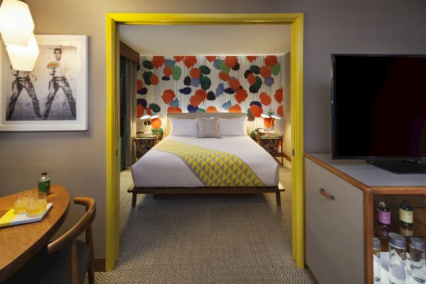 A cozy hotel room features a bed with colorful decor, patterned wallpaper, artwork, a TV, and a small table with drinks.