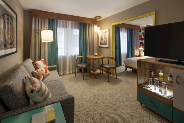 A cozy hotel room with a gray sofa, colorful cushions, round table with chairs, TV, and a bed in the background ends the sentence.