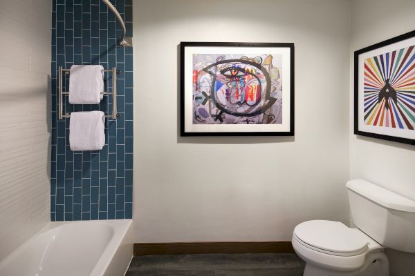 The image shows a bathroom with blue tile, a bathtub, art on walls, and a toilet.