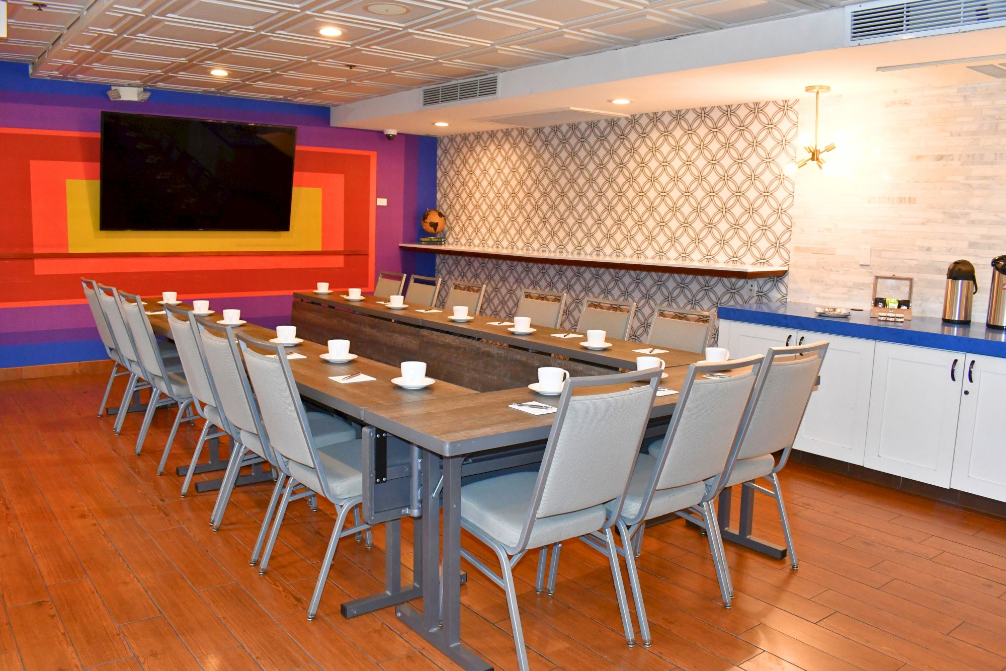 A conference room with a U-shaped table, set with cups, napkins, and cutlery. There’s a large TV, colorful wall art, and a refreshment area.