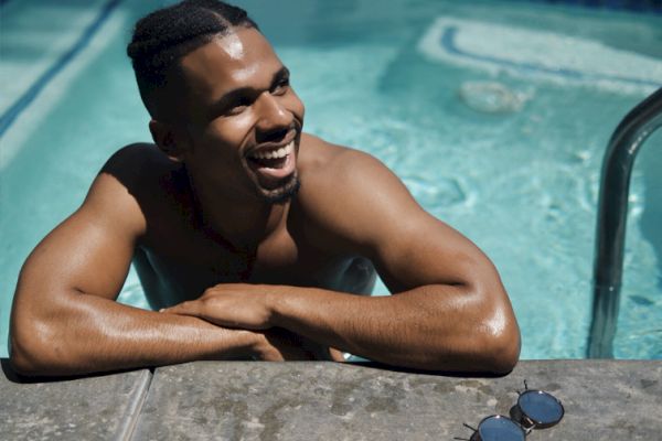 A person is smiling while leaning on the edge of a swimming pool, with sunglasses resting nearby on the poolside.