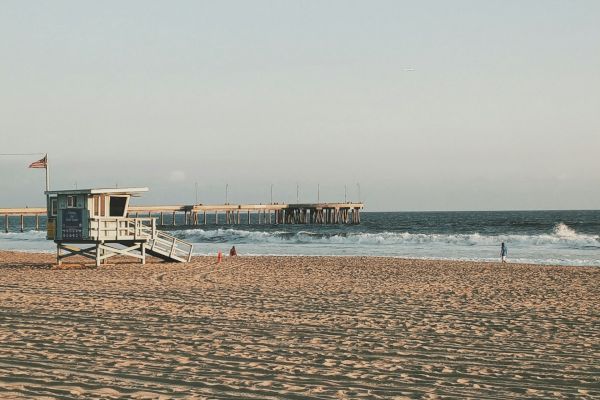 A beach scene with a lifeguard tower, people on the sand, a pier extending into the ocean, and a clear blue sky overhead.