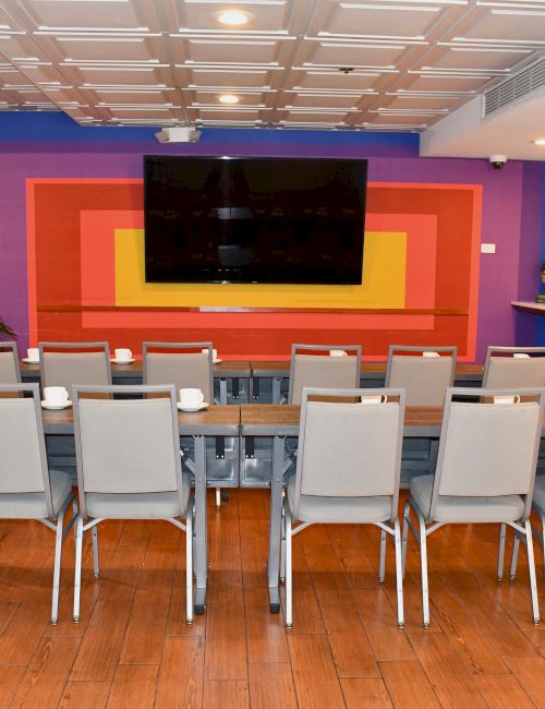 A conference room with a long table, chairs, a TV on a colorful wall, wooden floor, and exit sign.