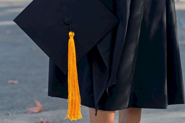 A person in a graduation gown holds a cap with a yellow tassel, wearing beige high heels.
