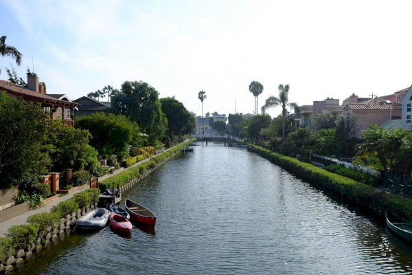 A serene canal lined with lush greenery and houses, small boats moored on the side, and palm trees under a clear, blue sky.