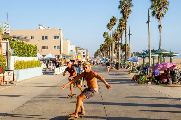 People are skateboarding on a beachside boardwalk with palm trees and various stalls in the background.